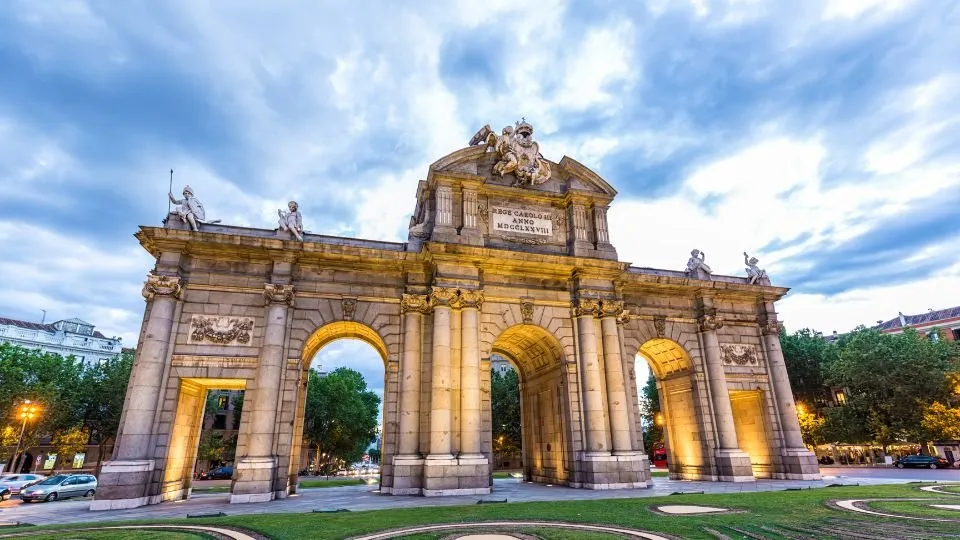 The Puerta de Alcalá at blue hour from below: a granite triumphal arch of five openings, three arched and two square, lit gold from within, with statues and a carved coat of arms on top under a cloudy sky