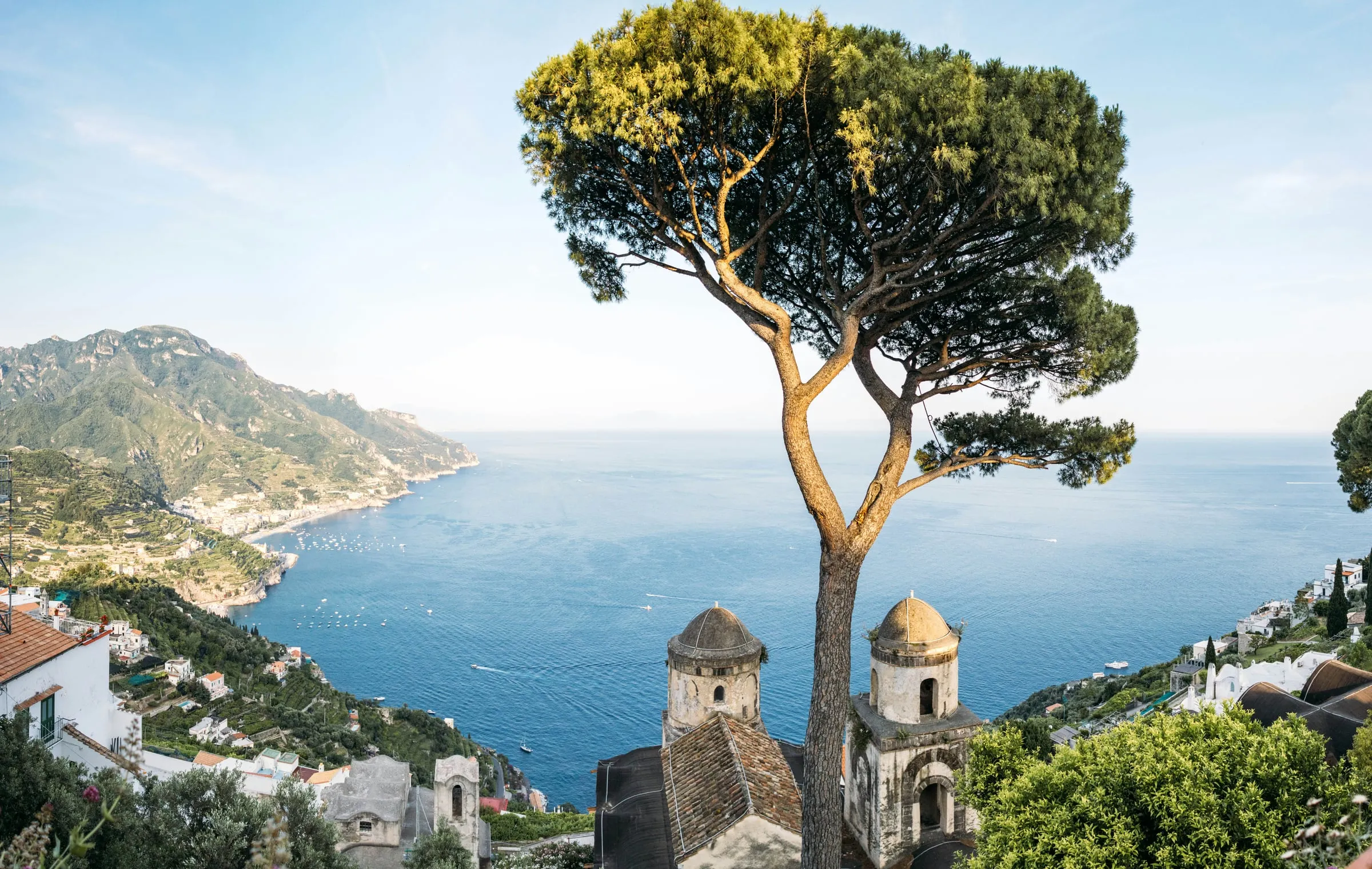 The view from Ravello: a tall umbrella pine on a terrace high above the sea, the two domed bell towers of a church below it, the coast running away to the left with a town on its shore, and mountains beyond under a pale sky