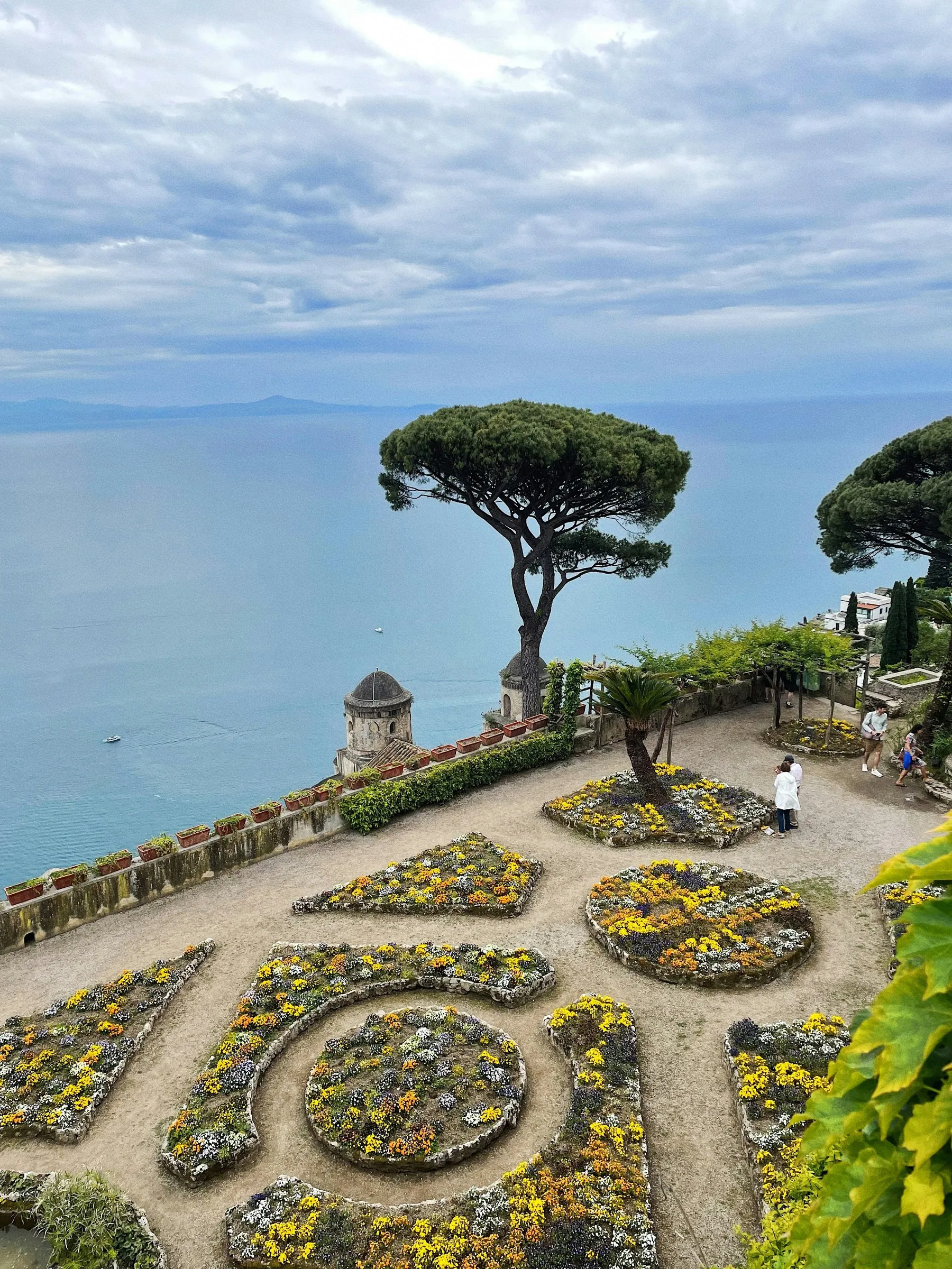 The gardens of Villa Rufolo in Ravello from above: geometric beds of yellow and white flowers on the belvedere terrace, a tall umbrella pine and a small domed bell tower at its edge, and the sea a long way below under a pale sky