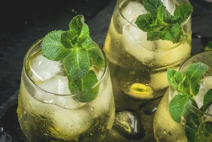 Three glasses of pale gold rebujito over ice, each topped with a sprig of mint, on a dark slate