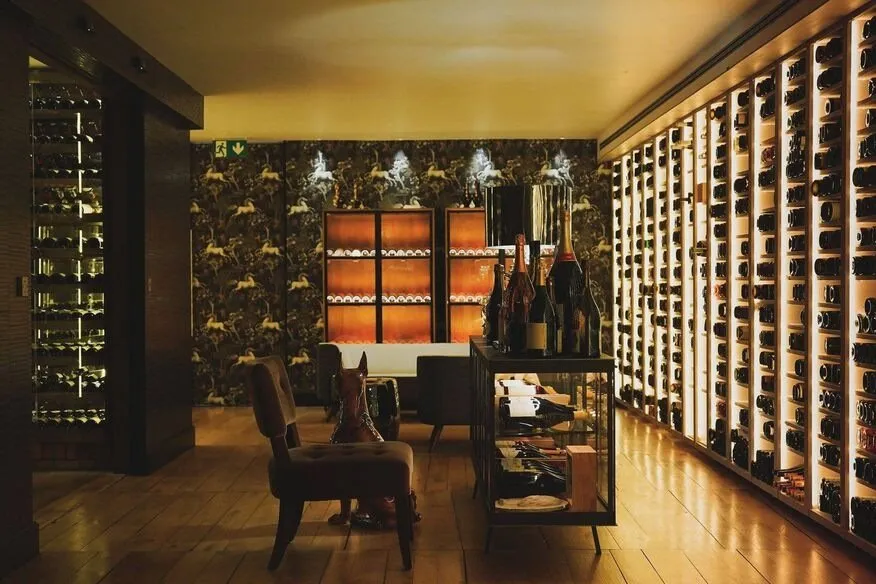 A wine room lit warm and low: a wall of back-lit racks holding bottles on the right, a glass case of bottles in the middle, an armchair and a dog sculpture on the wooden floor, and wallpaper of horses at the far end
