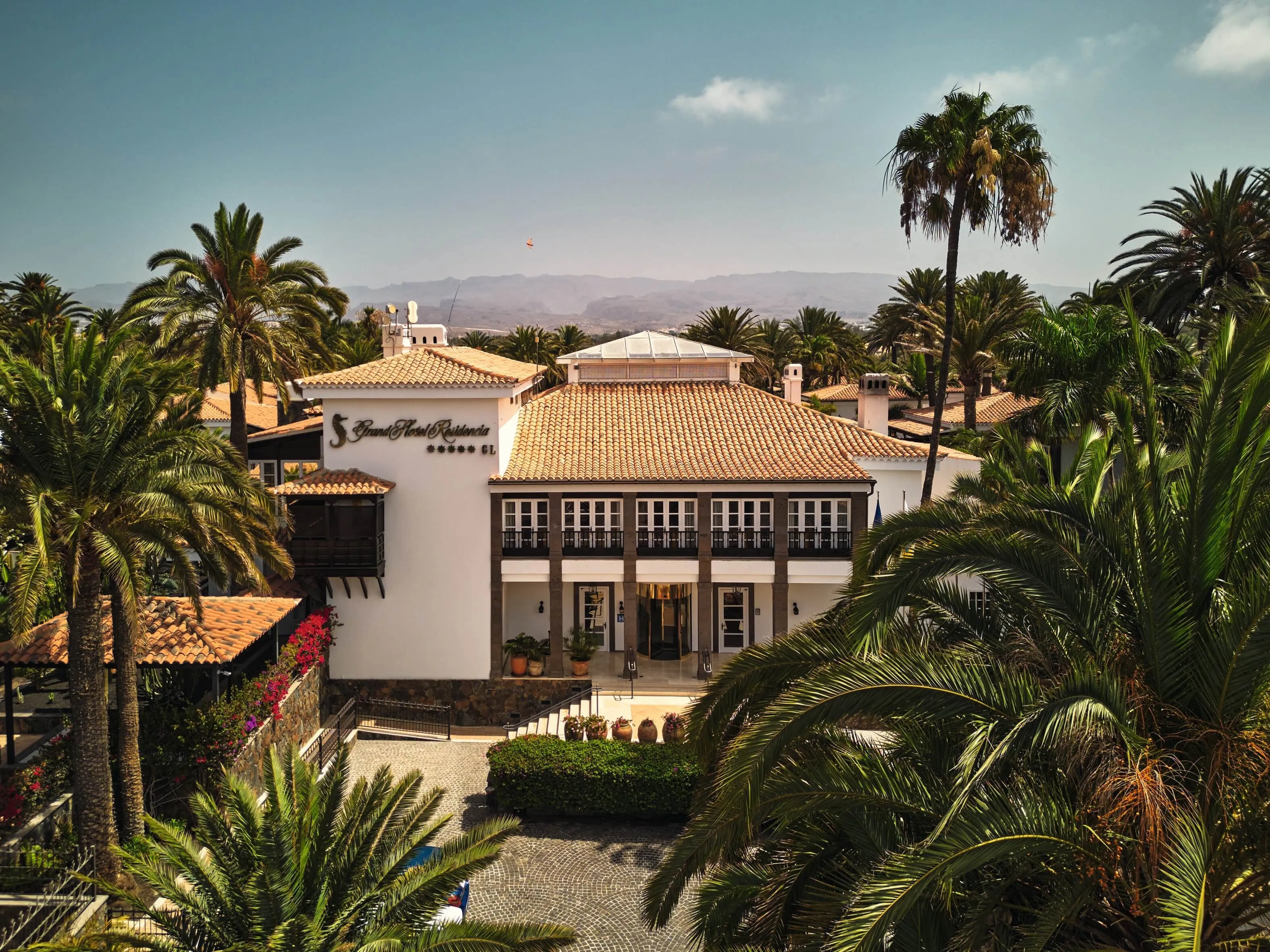 The hotel's main house seen from above across palm crowns: a white building with a terracotta roof, a glazed lantern, dark stone pillars and wooden balconies over the entrance, a paved forecourt, Gran Canaria's mountains in haze behind