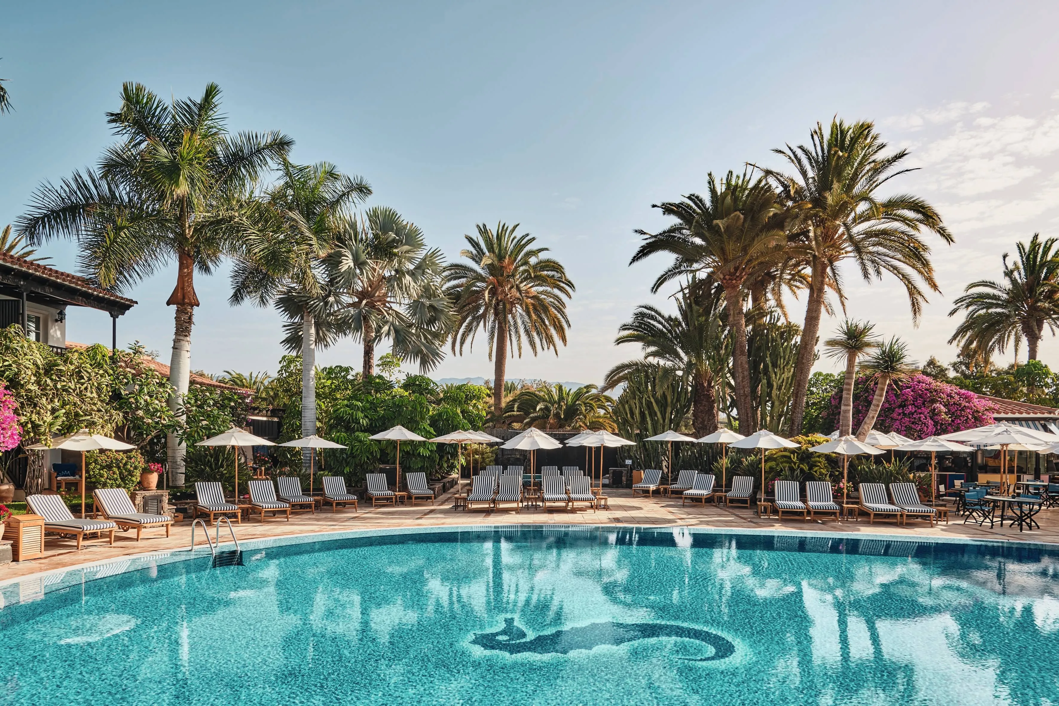The main pool, a curving turquoise basin with a seahorse in dark mosaic on its floor, rows of blue-and-white striped loungers and white parasols on the terrace beyond, tall palms and bougainvillea against a pale sky