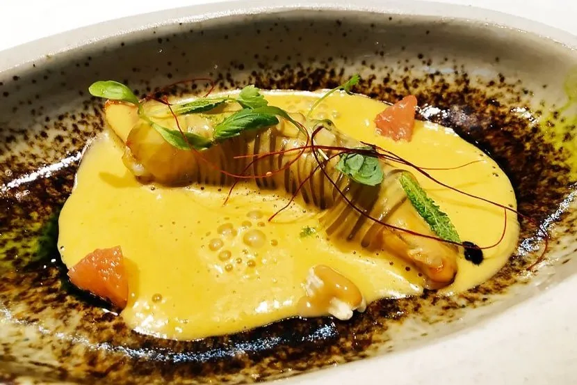 A ribbed pasta parcel and mussels in a pool of golden sauce, garnished with herbs and fine threads of red chilli, in a speckled stoneware dish