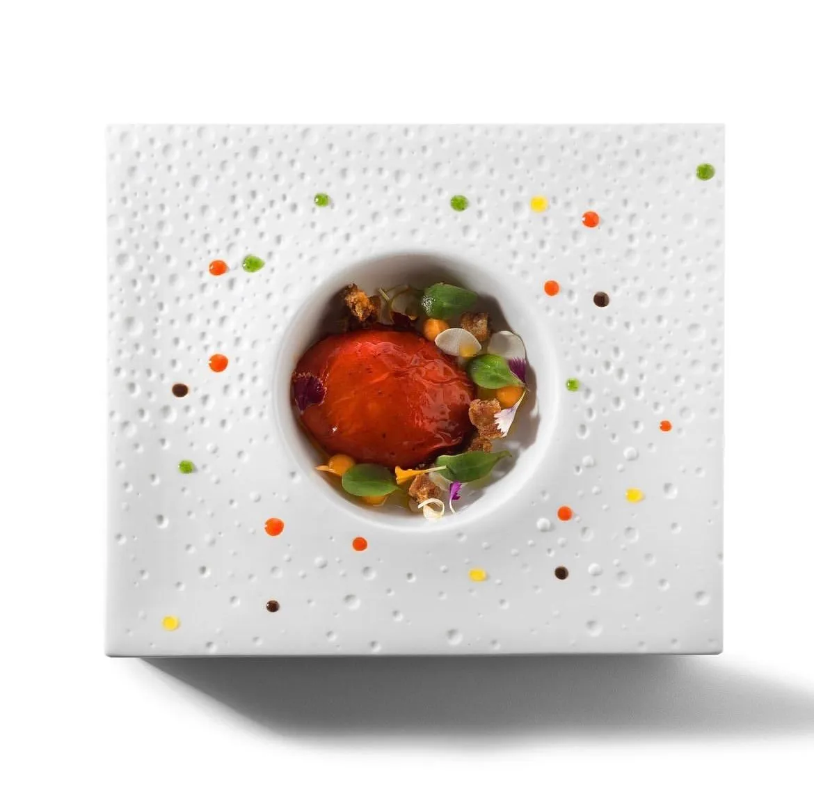 A white square plate dimpled like a sea sponge, a glazed red sphere in a well at its centre ringed with leaves, flowers and crumbs, and dots of coloured sauce scattered across the rim