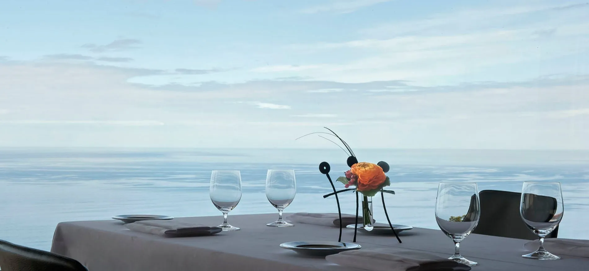 A table laid in grey linen with plates, folded napkins and wine glasses, an orange flower in a black wire stand at its centre, and a calm sea filling the window behind