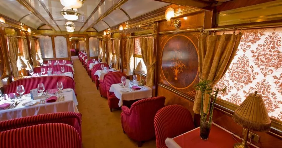A railway restaurant car with red striped armchairs at tables laid with white cloths and glasses, marquetry panels on the walls, damask-patterned blinds and glass lamps along a vaulted ceiling