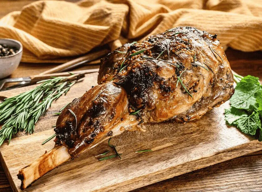 A roast leg of lamb on a wooden board, crusted with mustard seed and scattered with rosemary, sprigs of rosemary and fresh mint beside it and a yellow cloth behind