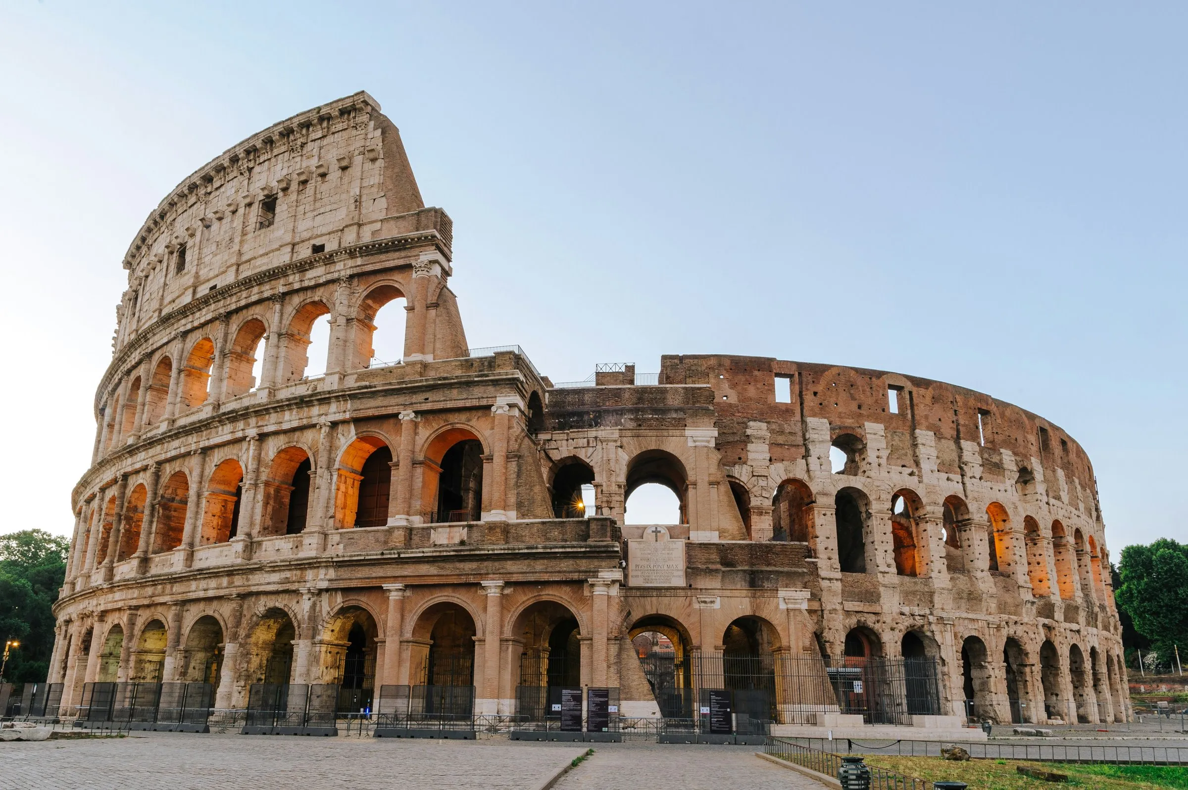 The Colosseum at sunrise seen from the west: the tall surviving side of the outer ring on the left with its inner arches lit orange, the lower ring curving away to the right, empty paving in front and a pale clear sky