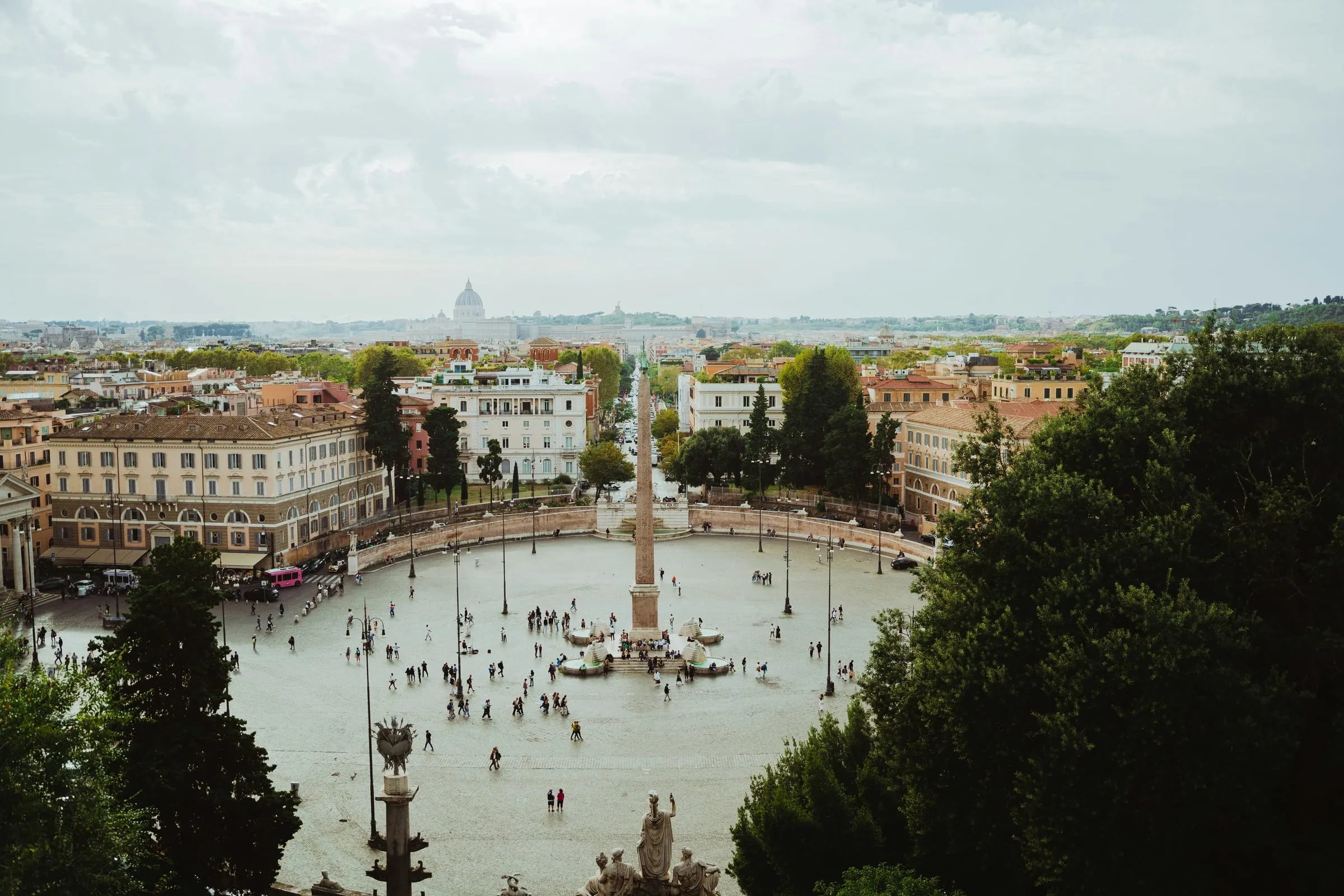 Piazza del Popolo seen from the Pincio terrace: the oval square with its obelisk and fountains, people crossing it as small figures, the streets of the centre beyond and the dome of St Peter's on the horizon under a soft grey sky