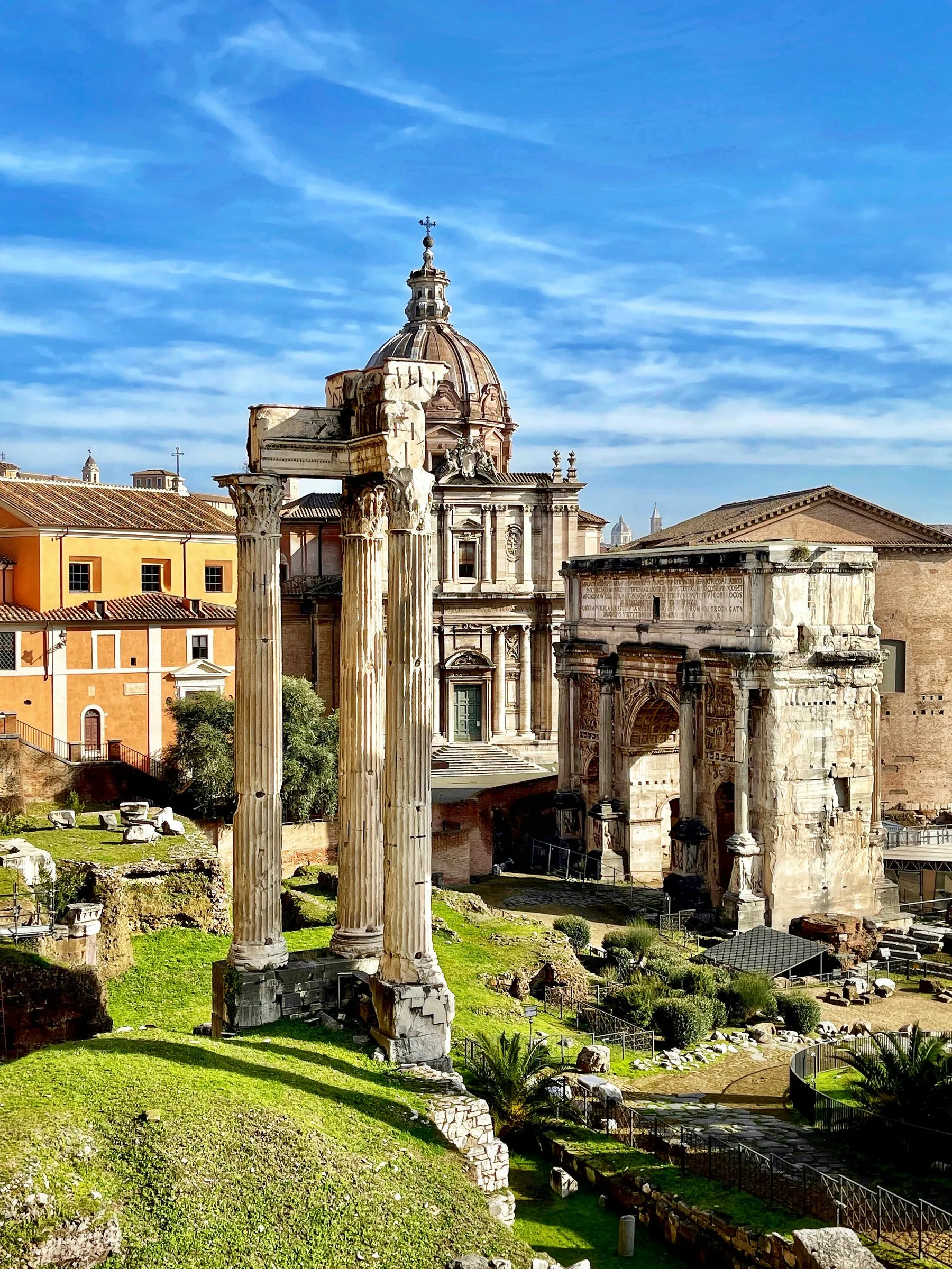 The Roman Forum under a blue sky with thin cloud: the three tall Corinthian columns of the Temple of Vespasian in the foreground, the domed church of Santi Luca e Martina behind them, the Arch of Septimius Severus to the right, and the grass and broken stones of the Forum below