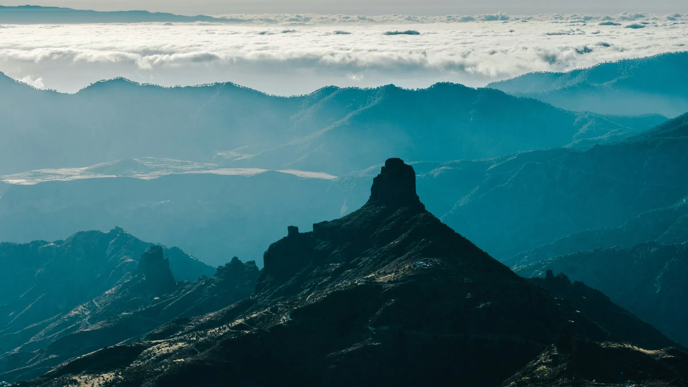 The dark rock peak of Roque Bentayga rising from a ridge, layers of blue mountains behind it and a sea of cloud on the horizon