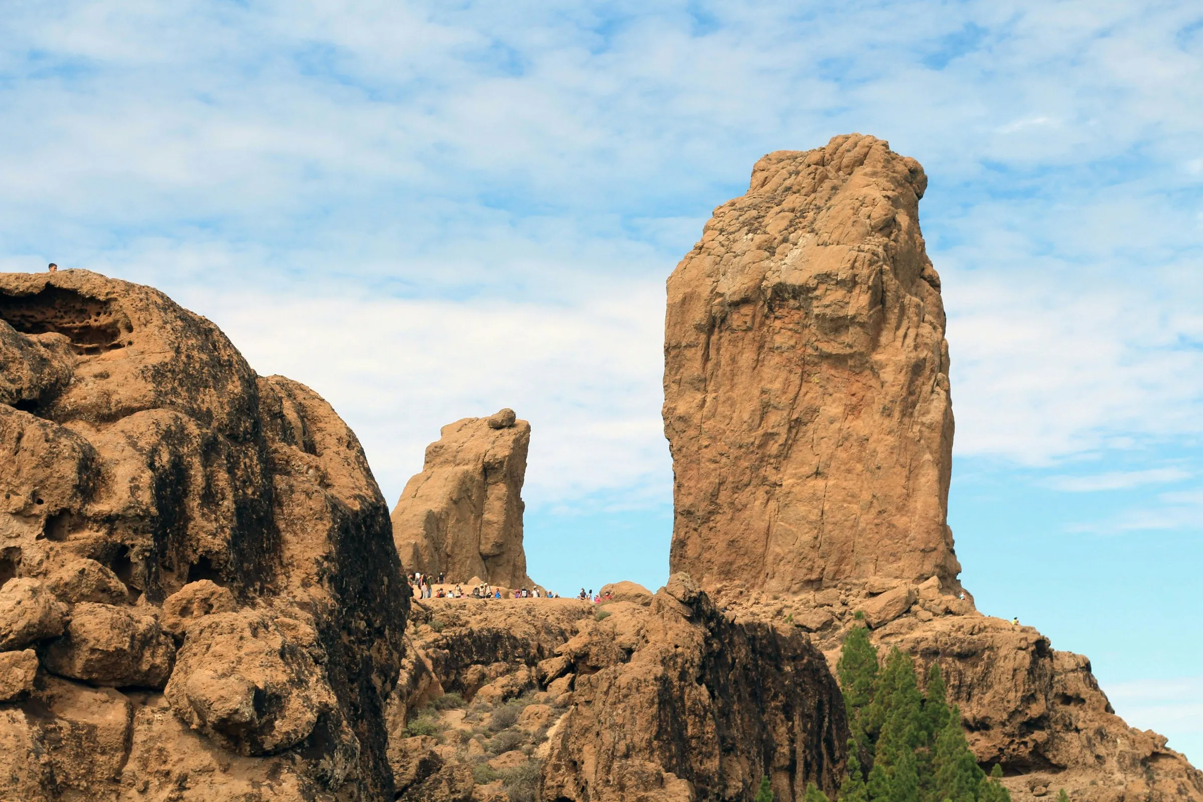 The tall ochre rock pillar of Roque Nublo on a rocky summit, a smaller rock beside it and boulders and pines in front, under a pale blue sky