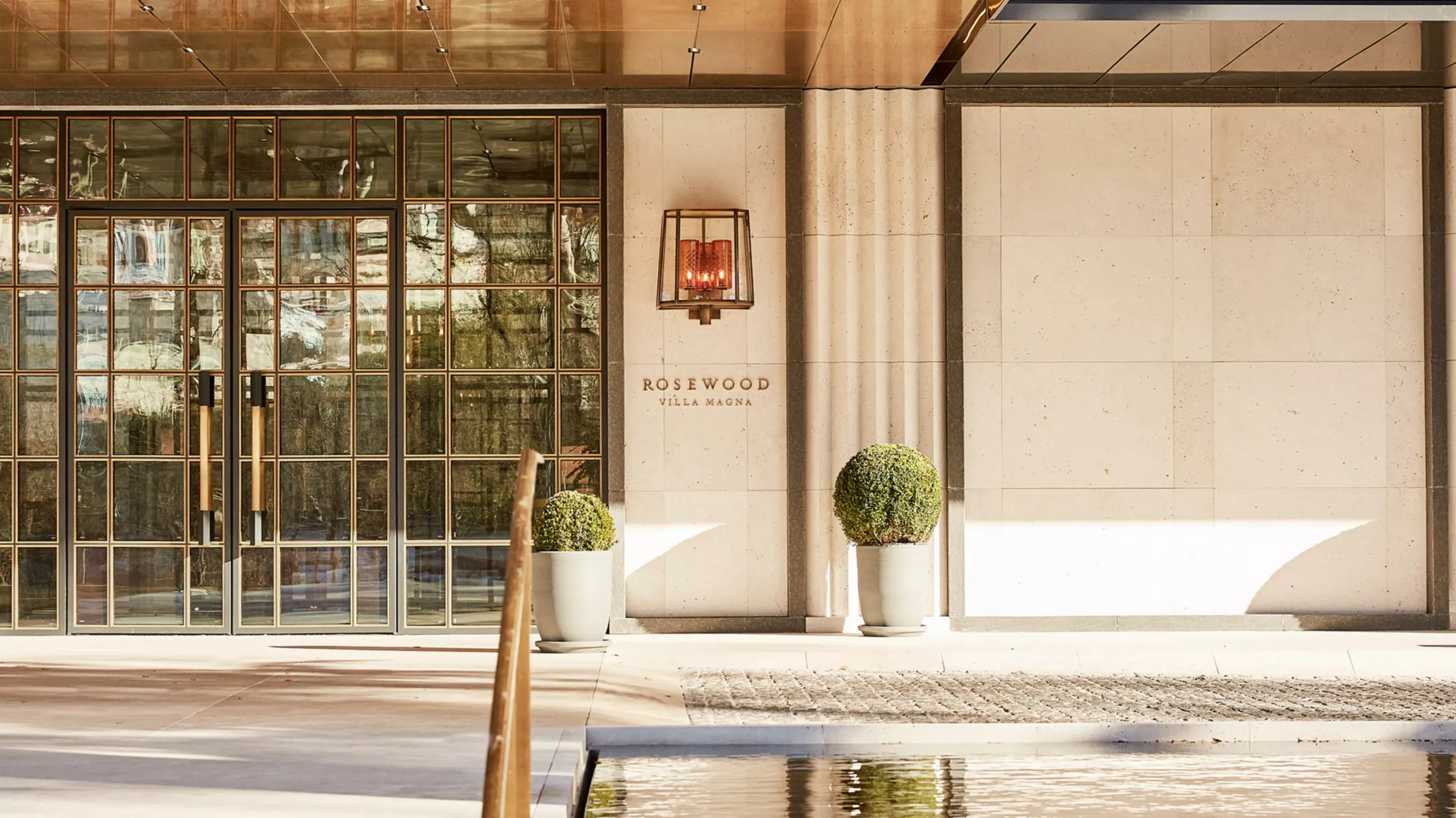The entrance of Rosewood Villa Magna: glass doors, a stone wall bearing its name, and a still pool in front