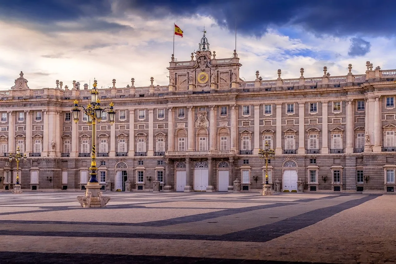 The Royal Palace of Madrid across its paved square: a long pale stone façade of columns and balustrades with a clock and the Spanish flag above the centre, a gilded lamp post on the left, under dark clouds