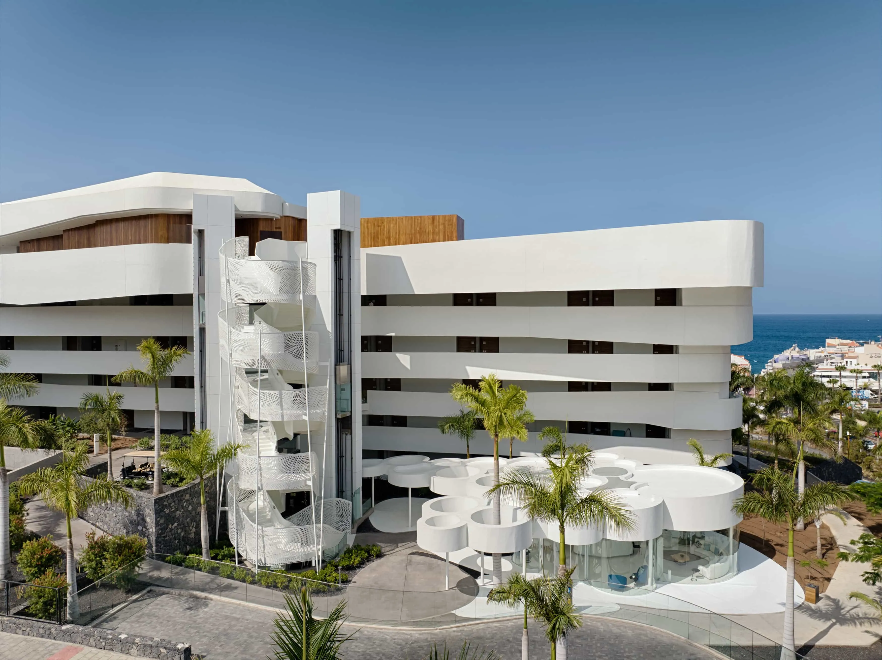 Royal Hideaway Corales Beach from above the street: a white building of long curved, stacked floors with dark timber bands, a white spiral staircase in a lattice drum at one corner, white disc-shaped pavilions on slender columns among palms in the forecourt, and the blue sea and the roofs of the coast beyond