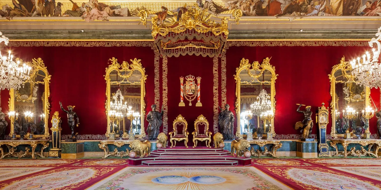 A throne room hung in red velvet: two gilded thrones on a red-carpeted dais beneath a gilt canopy, bronze lions at the steps, gilt mirrors and chandeliers along the walls and a painted ceiling above