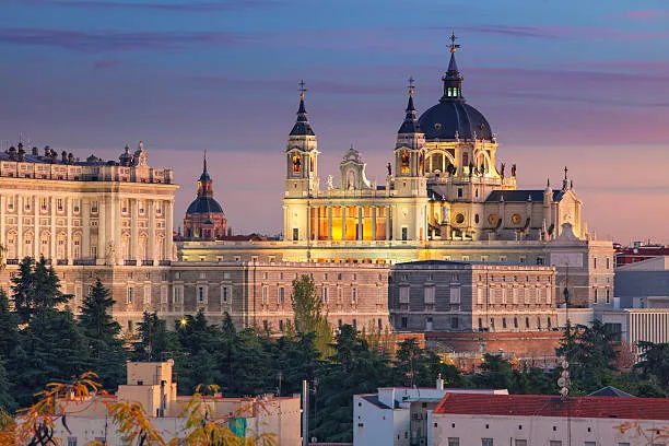Madrid at dusk: the dome and towers of the Almudena Cathedral lit gold, the long façade of the Royal Palace beside it, above dark trees and rooftops