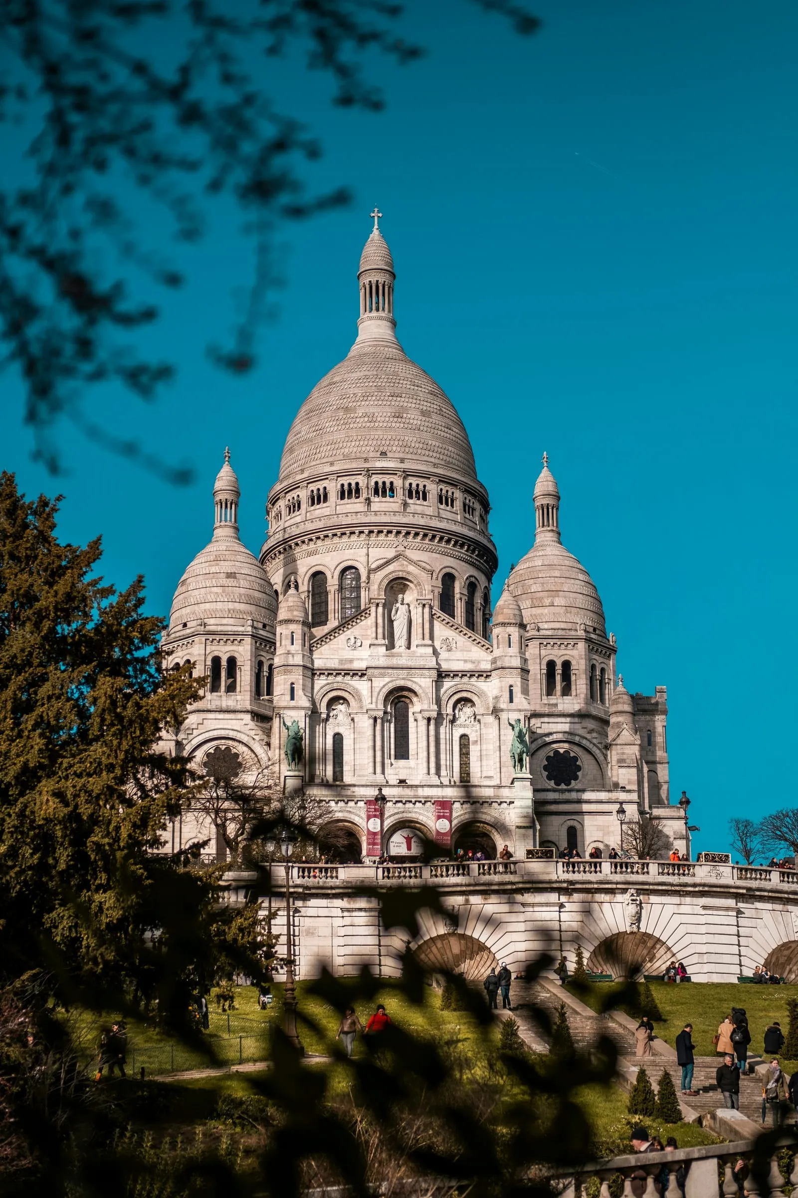 The white domes and towers of the Sacré-Cœur on a clear day, seen from the lawns below, bare branches blurred in the foreground and the double stairway rising to the terrace