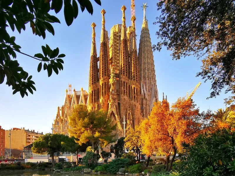 The spires of the Sagrada Família's Nativity façade glowing gold in low sun above a park of autumn trees, a crane beside the towers and a pond in the foreground