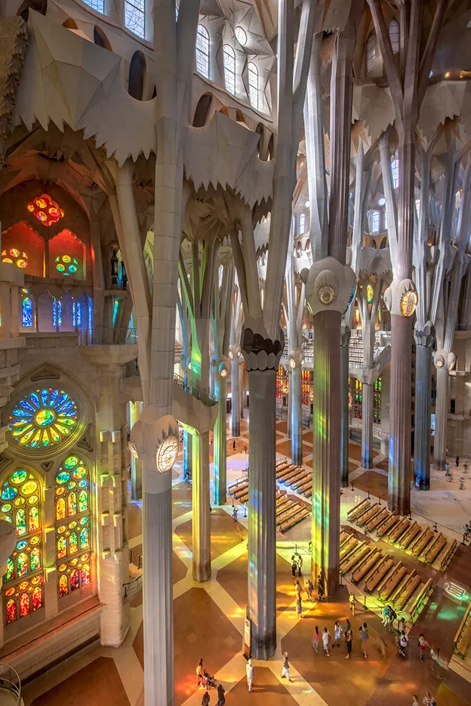 The nave of the Sagrada Família seen from a gallery: pale columns branching like trees into the vault, stained glass in the wall on the left throwing red, green and blue light across the floor and the rows of pews, a few visitors far below