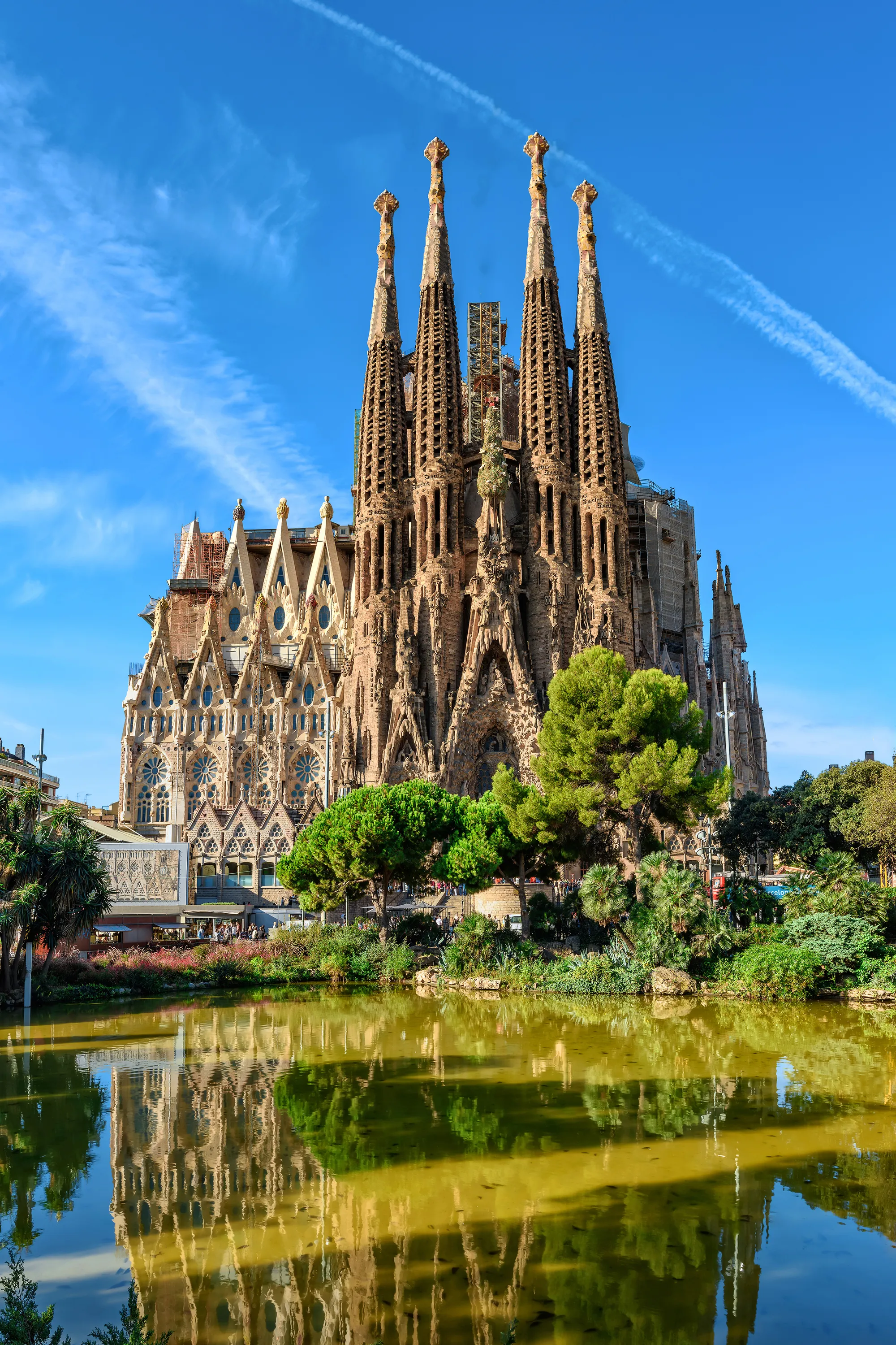 The Sagrada Família's Nativity façade, four tall carved spires above its portals and trees, mirrored in a still green pond under a blue sky streaked with cloud