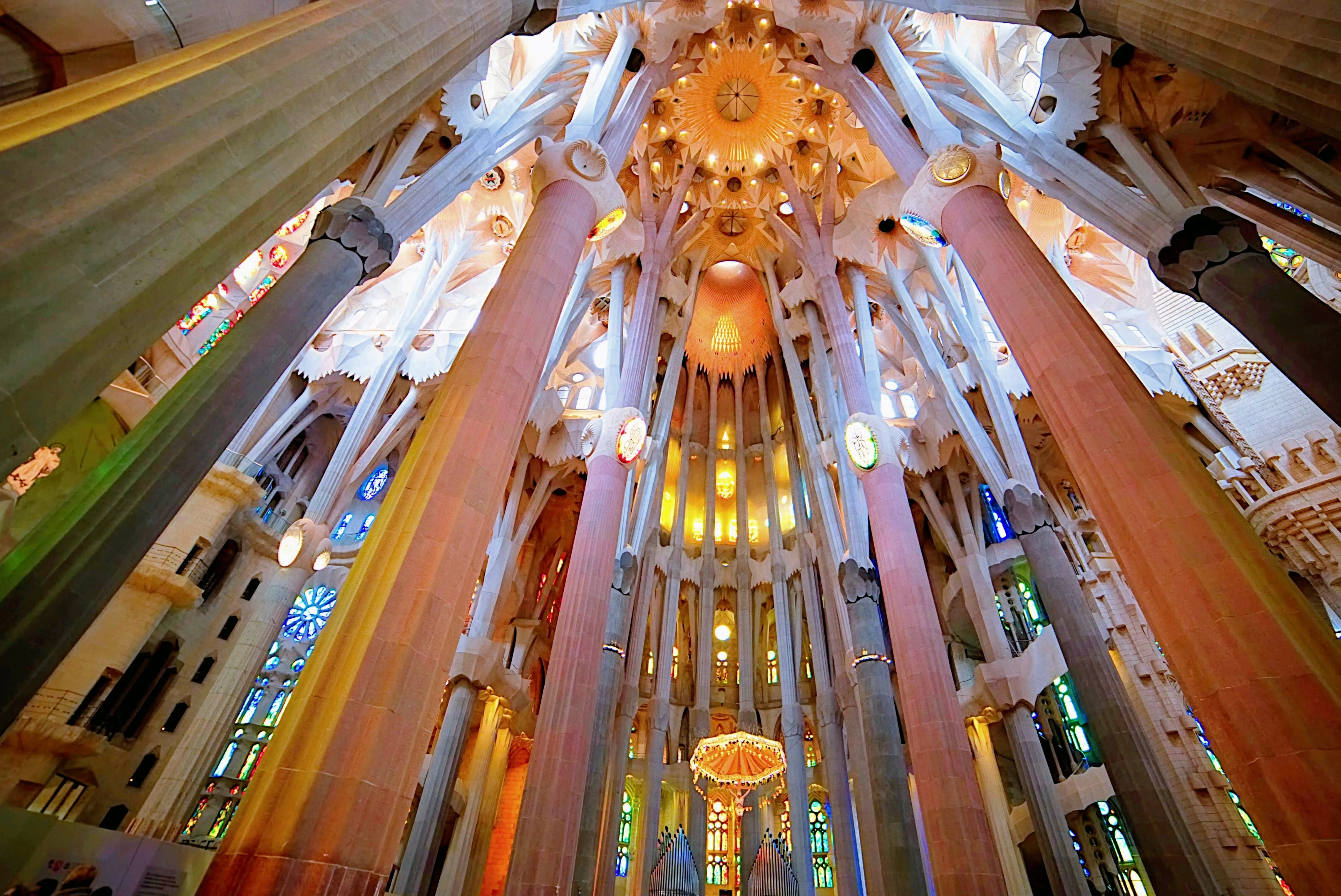 The nave of the Sagrada Família: tall columns branching like trees into a white and gold vault, lit orange, green and blue through the stained glass