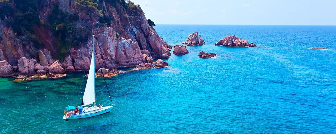 A white sailing boat with its mainsail up in clear turquoise water below pink granite cliffs and pines, rocky islets rising from the sea beyond and the open Mediterranean to the horizon
