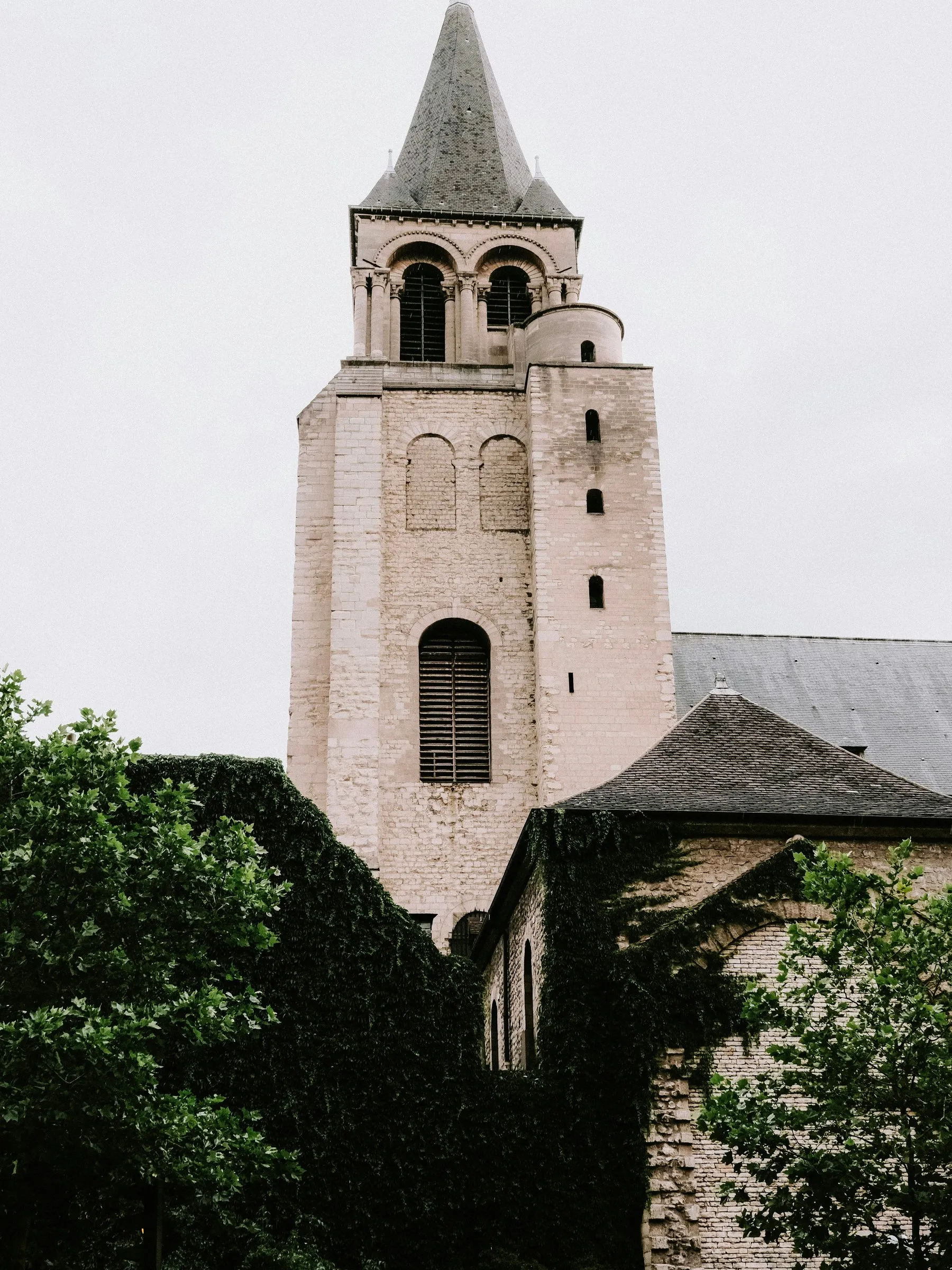 The bell tower of Saint-Germain-des-Prés on a grey day: a square Romanesque tower of pale stone with paired belfry openings under a slate spire, a round stair turret against its side, the nave roof and an ivy-covered wall below