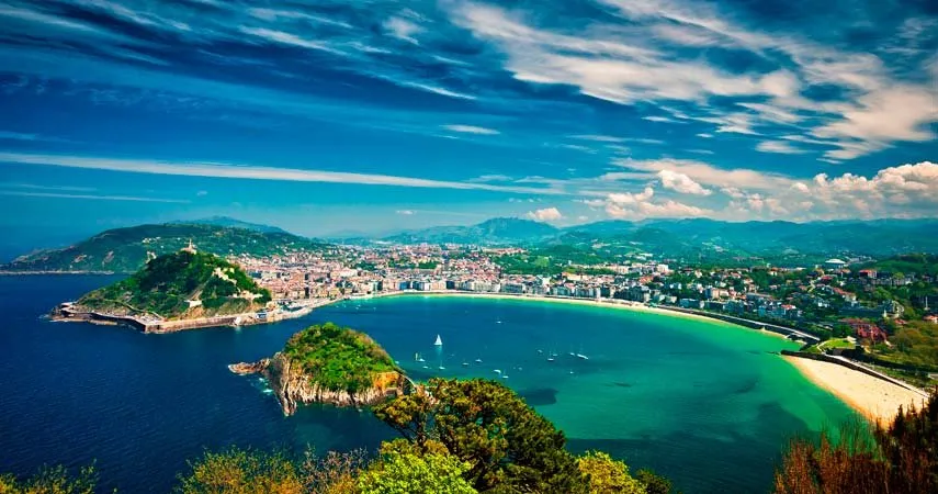 A wide bay seen from a wooded hilltop: a green island with rocky cliffs in turquoise water, sailing boats at anchor, a long curve of sand and the city behind, a second island-like hill on the left and mountains under a streaked blue sky