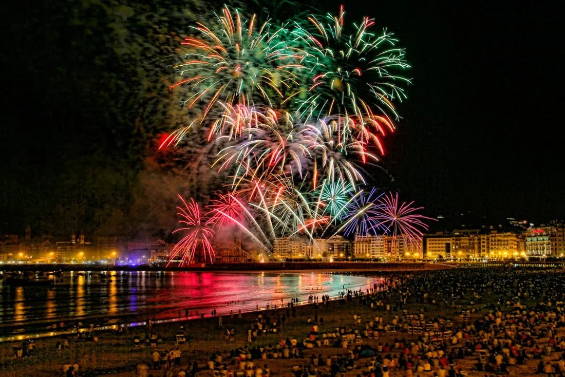Fireworks bursting green, gold and red over La Concha Bay at night, their colours mirrored in the water, with crowds sitting on the beach and the lit buildings of the promenade beyond