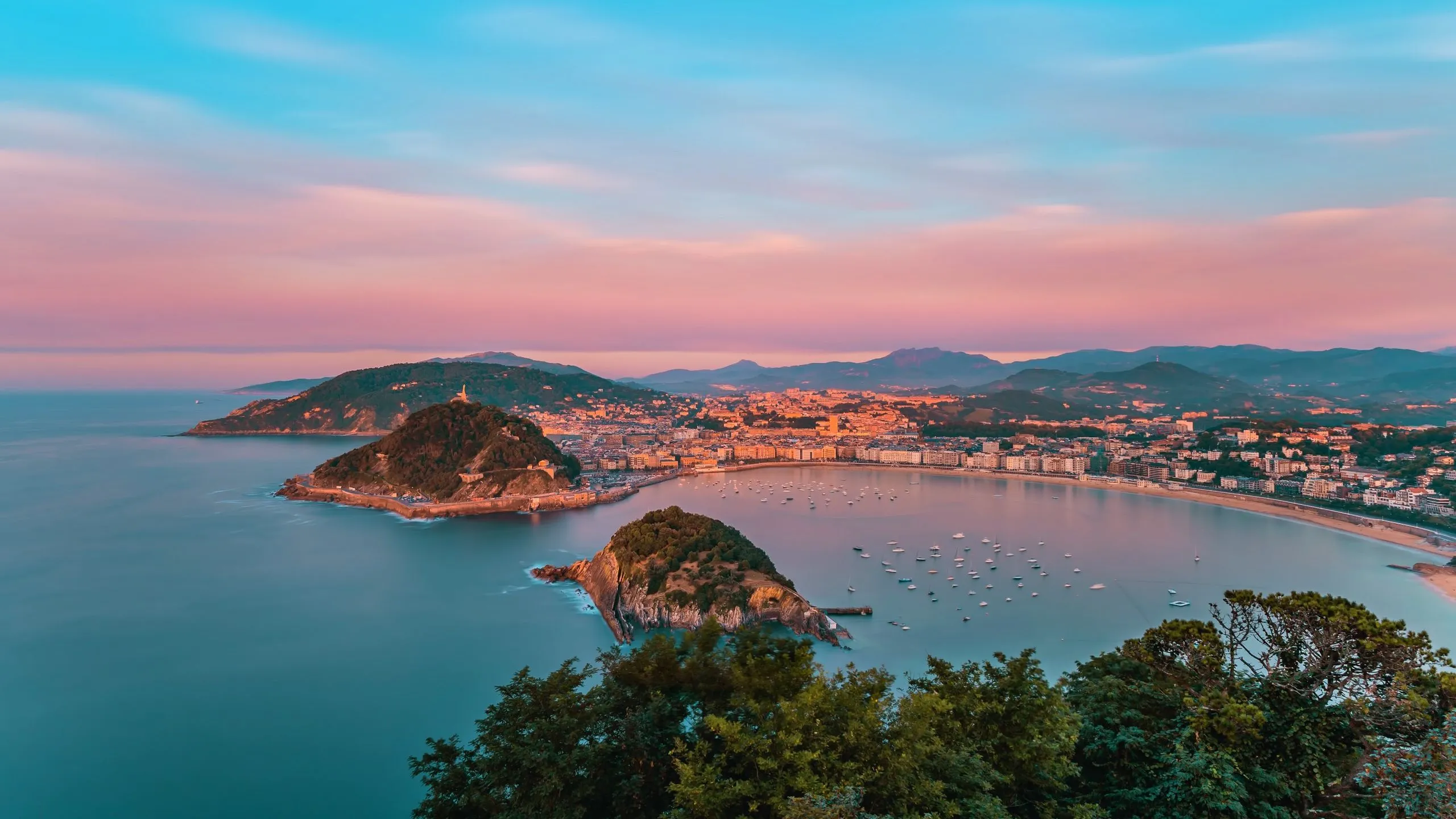 La Concha Bay at dusk from high above: the wooded island of Santa Clara in the bay, Monte Urgull behind it with a statue on its summit, and the city curving along the beach under a pink sky