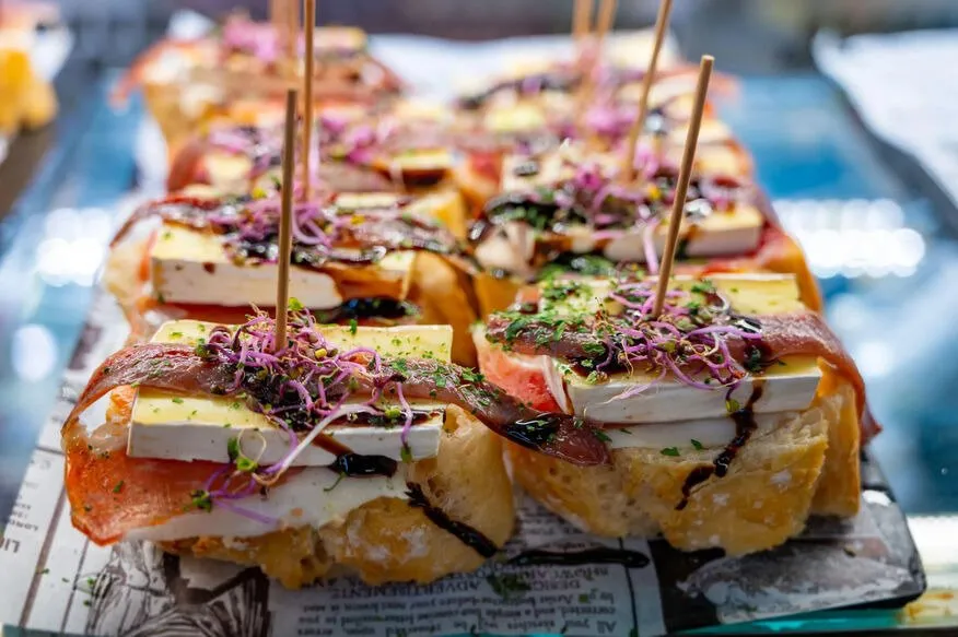 Pintxos on slices of bread laid on newspaper: cured ham and soft cheese topped with purple shoots, chopped herbs and a dark glaze, each held with a wooden stick