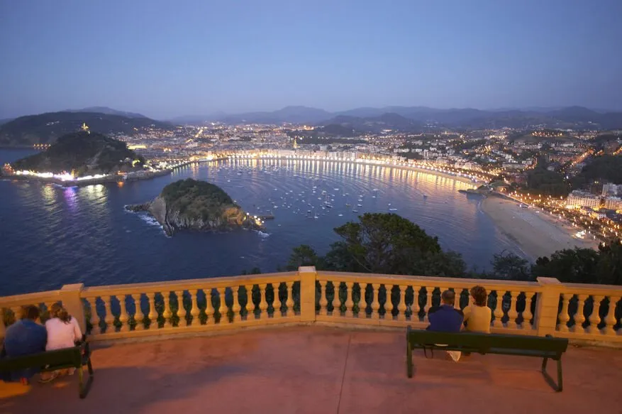 The bay of La Concha at dusk from a balustraded terrace high above it: the curve of lit beach and promenade, an island in the bay and the city's lights under the hills, two couples on benches at the rail