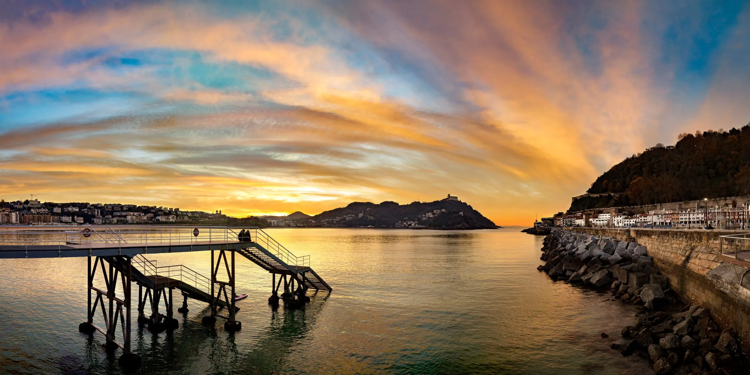 Sunset over La Concha Bay under streaks of orange and blue cloud: a steel platform with stairs stands in the water at the left, Monte Igeldo rises on the skyline, and the wooded slope of Monte Urgull and the harbour's houses line the sea wall at the right