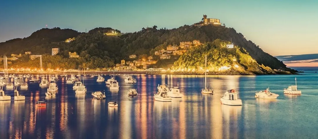 Boats moored in La Concha Bay at dusk, their lights reflected in the still water, beneath the wooded slopes of Monte Igeldo, lit along the shore and crowned by a castle-like tower at its summit