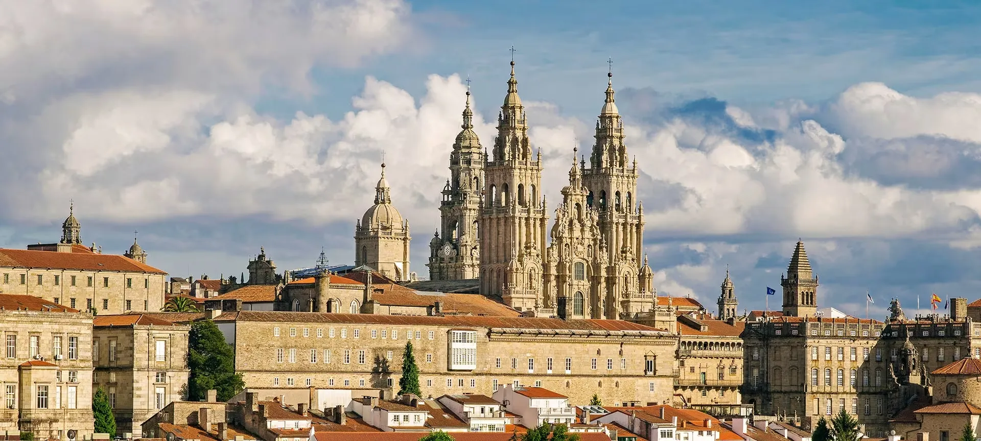 The baroque towers of Santiago's Cathedral rising above the old town's long granite buildings and red-tiled roofs, under a blue sky heaped with white cloud