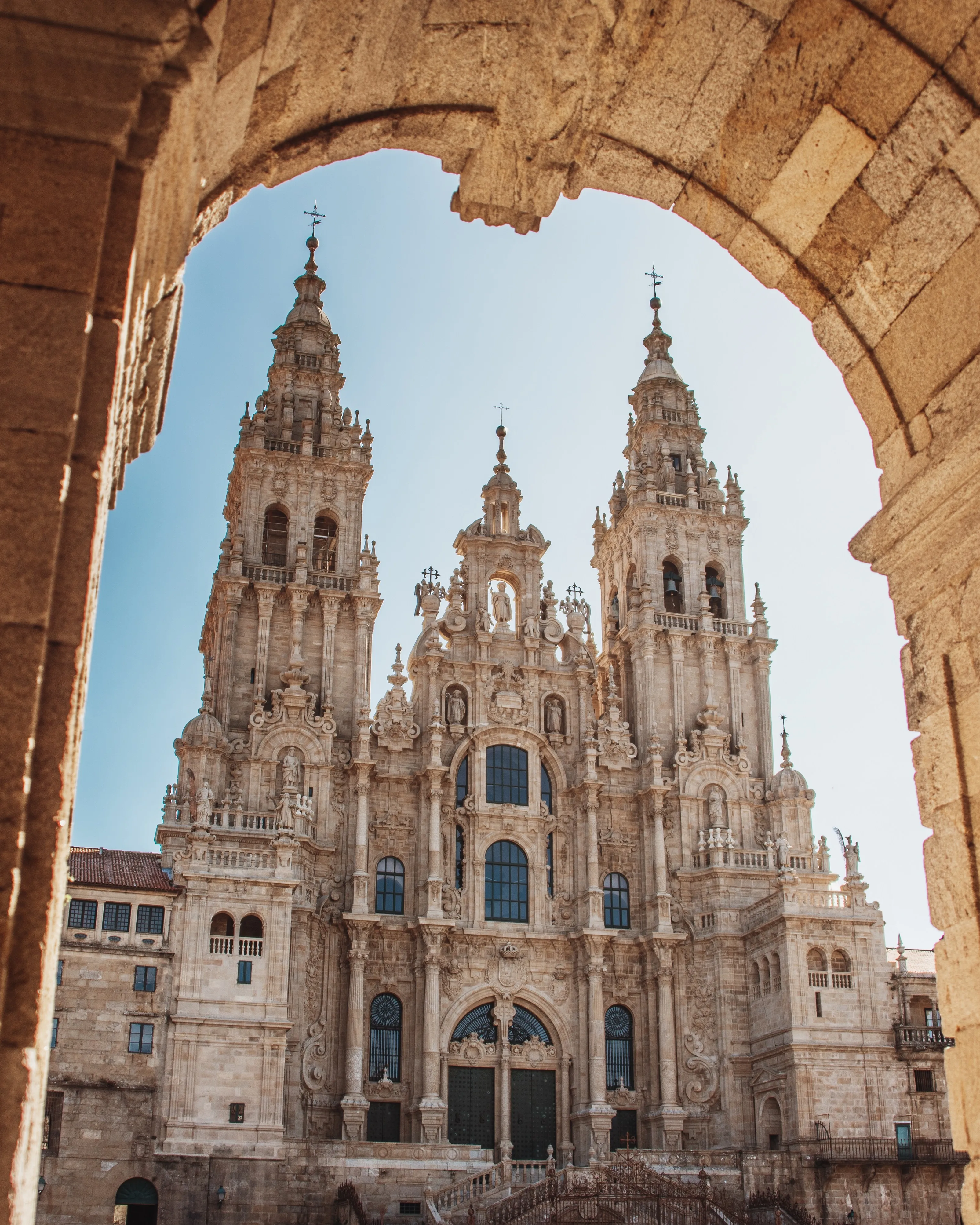 The Obradoiro façade of Santiago's Cathedral framed by a dark stone arch: two tall Baroque towers and a central gable carved with statues, the granite warm in the sun against a pale blue sky