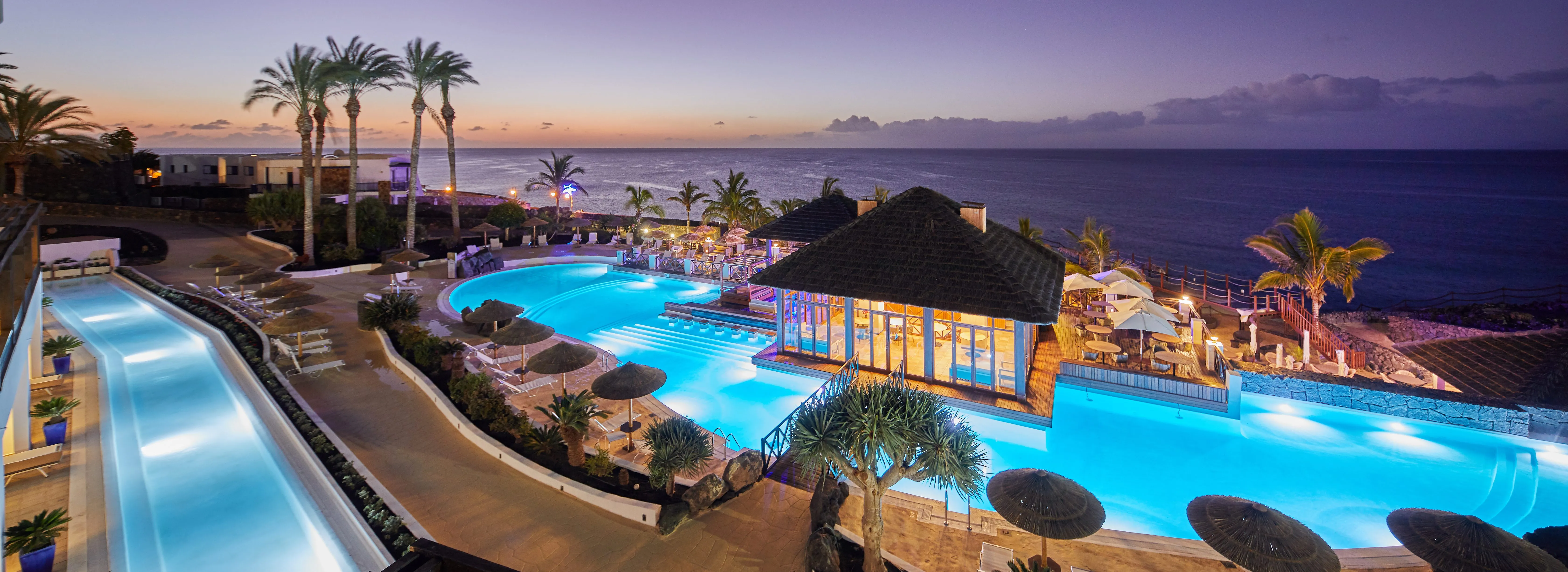 The pools at Secrets Lanzarote lit at dusk, a thatched pavilion between them and the Atlantic beyond under a violet sky