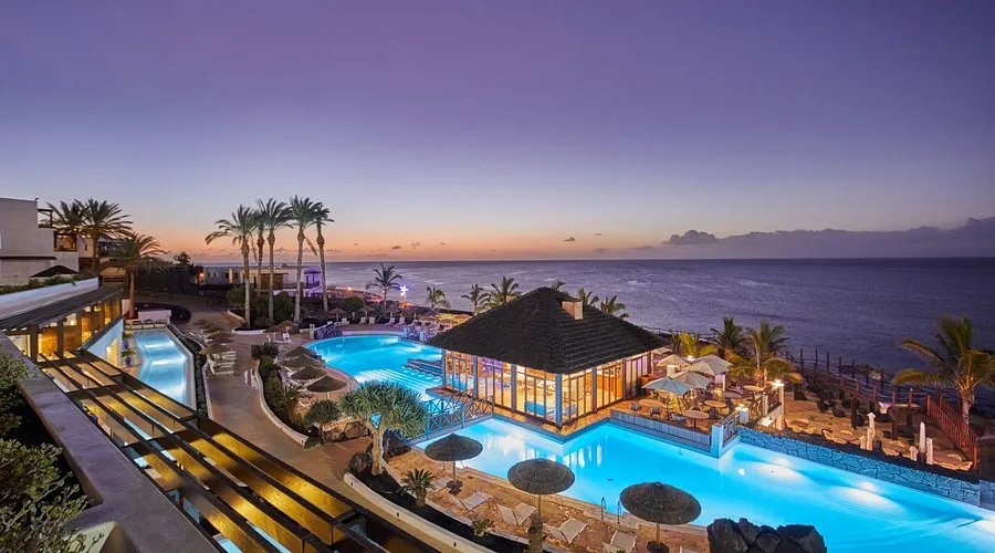 The pools at Secrets Lanzarote at sundown, a lit thatched pavilion between them and the sea beyond