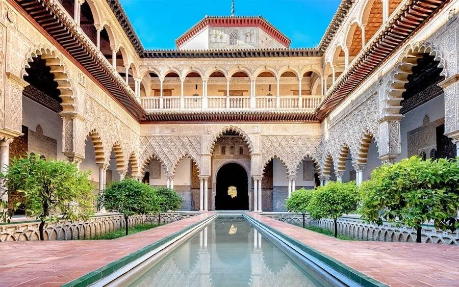 The Patio de las Doncellas in the Alcázar of Seville: carved stucco arches around a long reflecting pool, with orange trees in sunken gardens on either side
