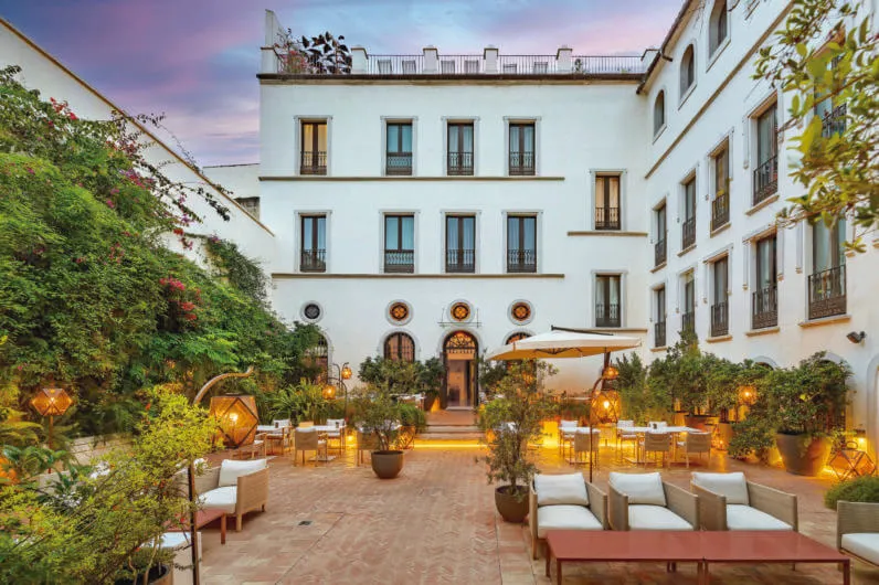 The hotel's patio at dusk under a pink sky, with lanterns lit among orange trees and bougainvillea, sofas and laid tables on herringbone brick below a white four-storey façade