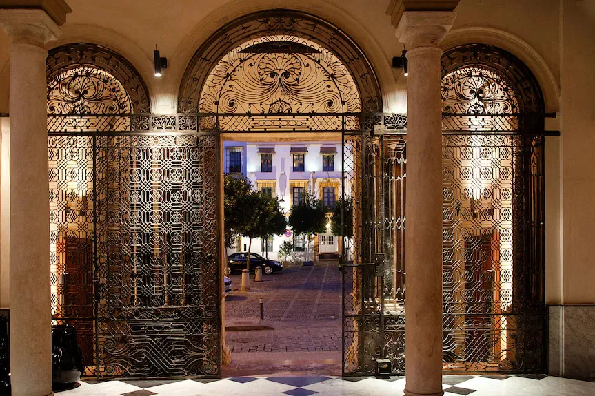 The palace entrance at night, looking out through tall wrought-iron gates with scrolled overpanels to an orange tree and a lit façade across the street