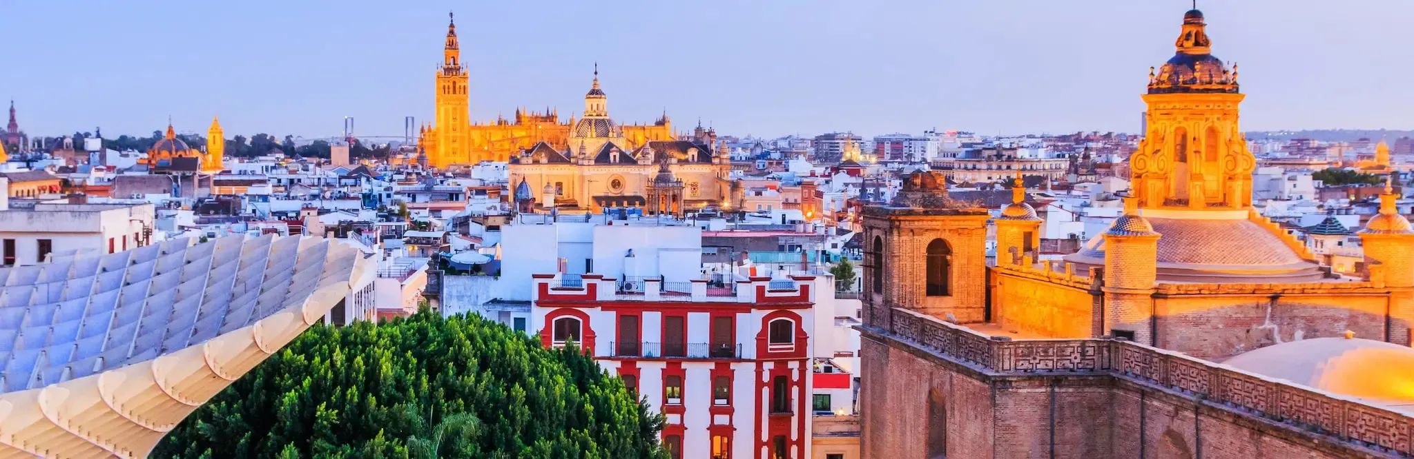 Seville's rooftops at dusk from the Metropol Parasol, its timber canopy in the foreground, the Giralda and the Cathedral lit gold on the skyline and a floodlit domed church to the right