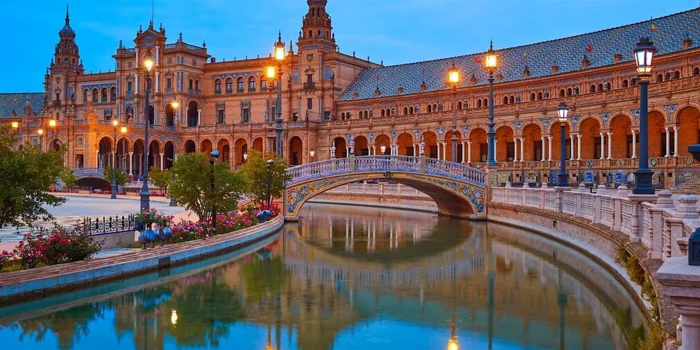 The Plaza de España in Seville at dusk, its lamps lit and a tiled bridge reflected in the canal