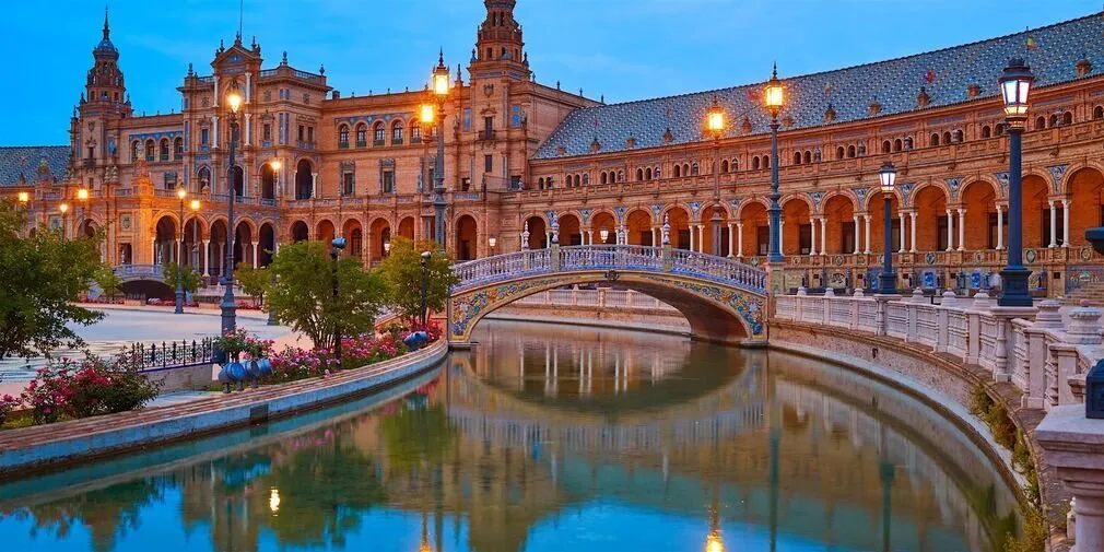 The Plaza de España in Seville at dusk, its lamps lit and a tiled bridge reflected in the canal