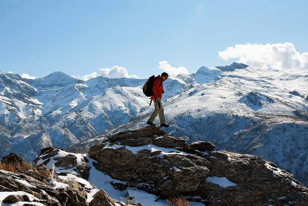 A walker in a red jacket and rucksack crossing a rocky outcrop dusted with snow, the snow-covered ridges of the Sierra Nevada behind under a blue sky