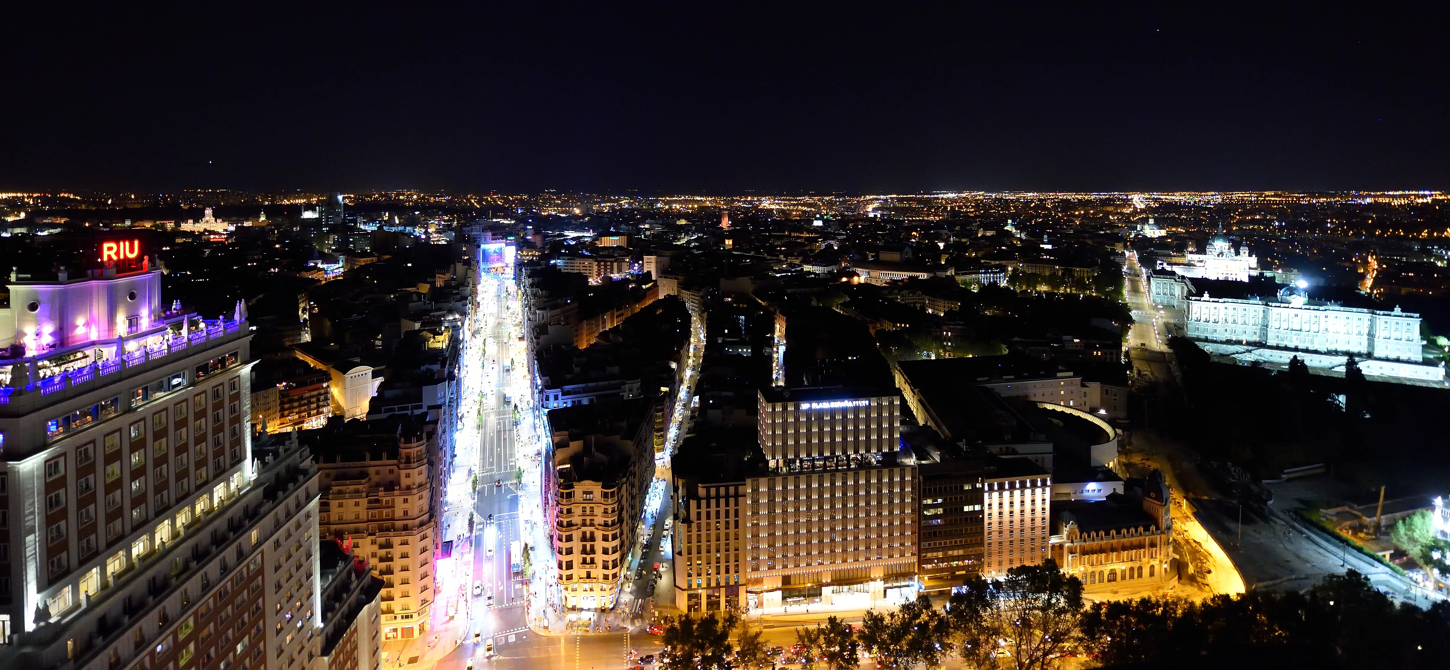 Madrid at night from high above: a lit street running away between the city blocks, the Hotel Riu Plaza España with its red sign on the left, and the floodlit Royal Palace on the right with the Almudena Cathedral behind it
