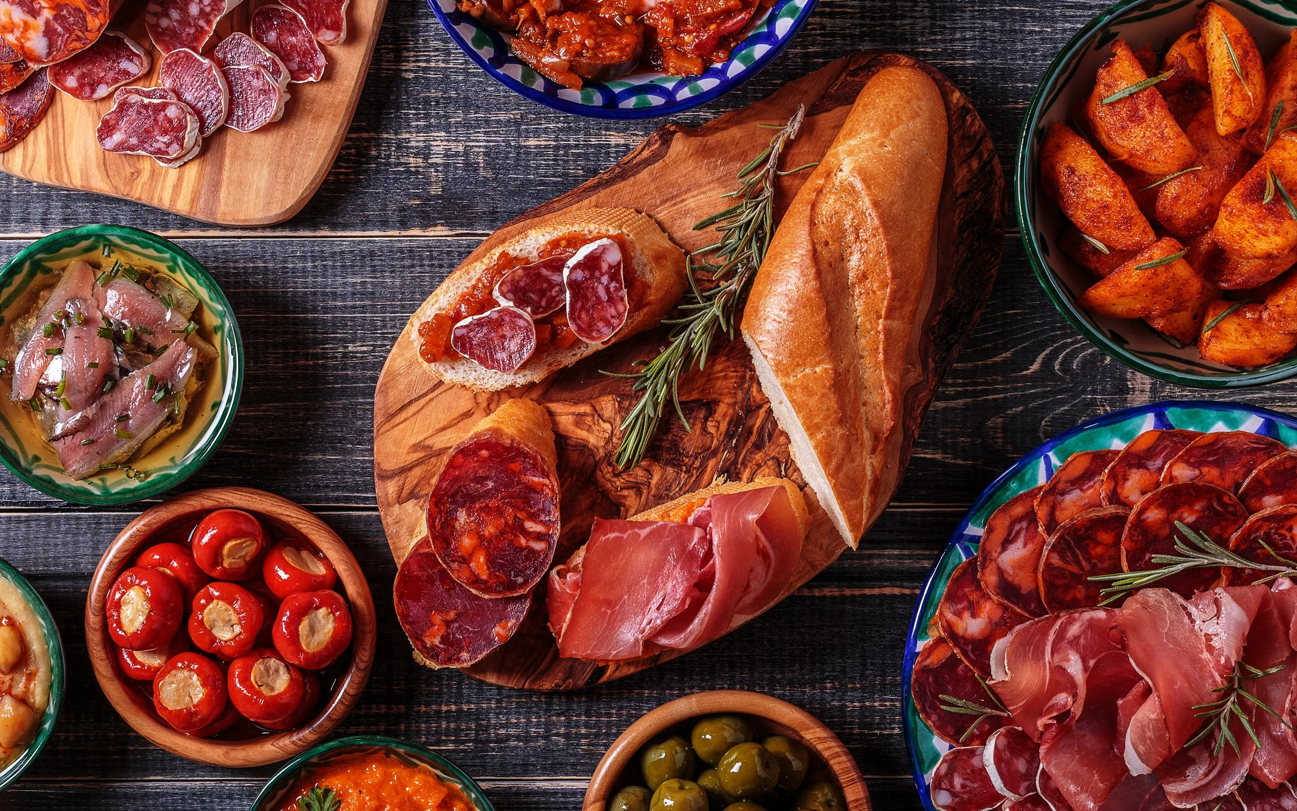 A tapas spread from above on dark wood: a baguette on an olive-wood board with slices of jamón and chorizo, patatas bravas, stuffed red peppers, anchovies and a bowl of olives