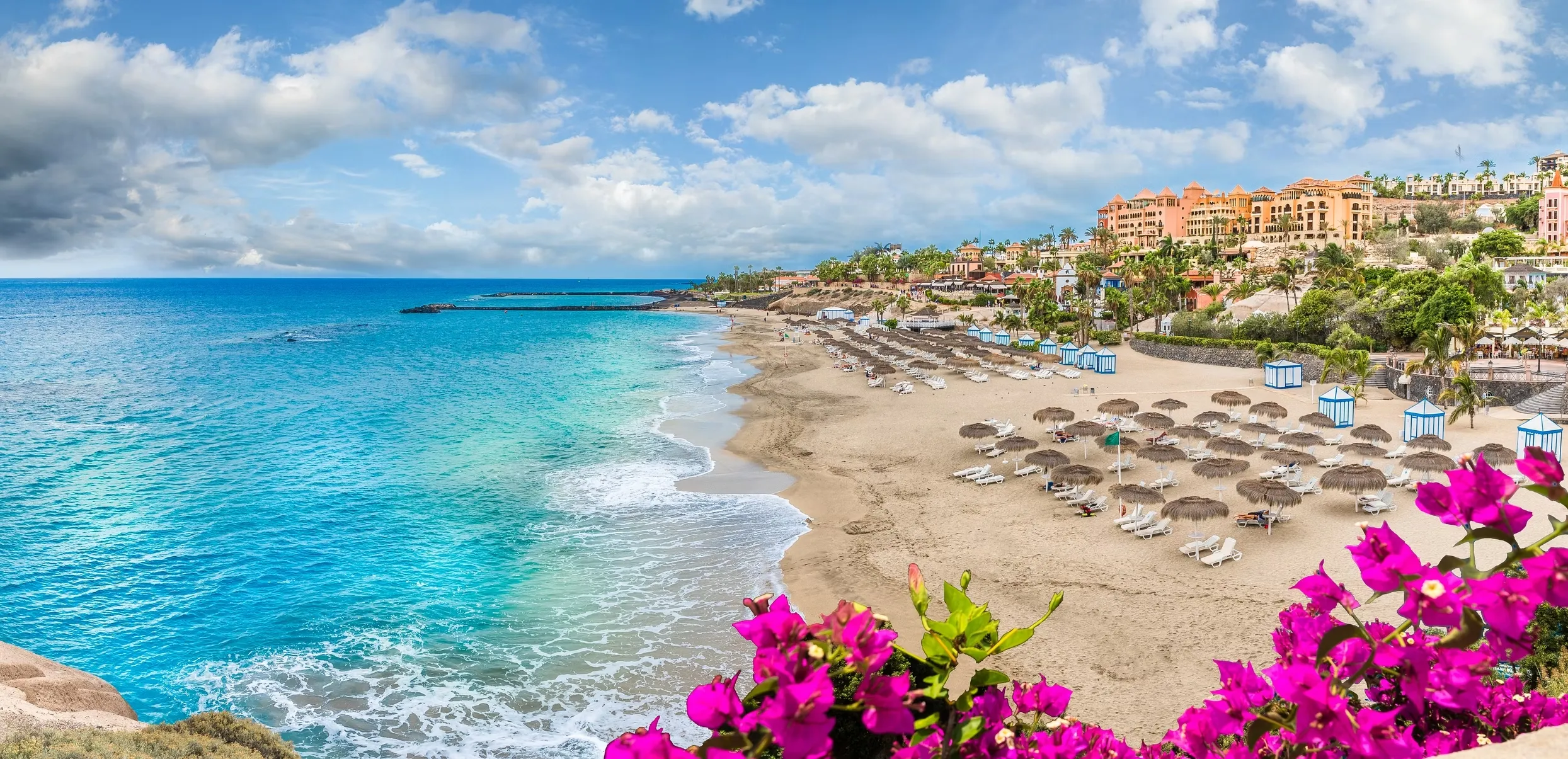 A curve of pale sand on the Costa Adeje: straw parasols and blue-and-white beach huts on the sand, a terracotta resort on the headland behind, bougainvillea in the foreground